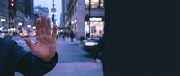Person with hand raised in stop gesture on urban street at dusk, wearing dark jacket. Suitable for concepts of refusal, rejection, traffic safety, and city life communications.