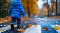 Young child in blue winter jacket walking alone on wet autumn sidewalk with fallen orange leaves, creating nostalgic mood in urban park setting.