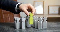 Business hand selecting green figure from group of gray people icons on office desk representing recruitment, leadership choice, and human resources talent selection.