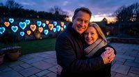 Happy mature couple embracing outdoors at twilight with colorful heart-shaped bokeh lights creating romantic atmosphere for Valentine's Day celebration.