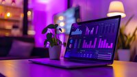 Laptop displaying financial analytics dashboard with purple ambient lighting and decorative plant, showing business data charts and graphs in dark mode.