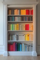 Books arranged by color spectrum on white built-in bookshelf with classic molding, creating rainbow effect against neutral wall, modern interior design element.