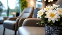 Fresh white daisies in decorative vase on armrest of brown leather chair, modern waiting room interior with bokeh lights and window view background.