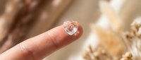 Small transparent heart-shaped crystal or gemstone balanced on fingertip against warm golden bokeh background for jewelry design and romantic concepts.