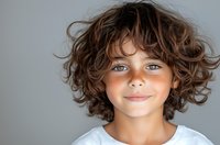 Young person with bright blue eyes and natural curly brown hair wearing white t-shirt smiling warmly against neutral gray background. Genuine expression of joy and innocence.