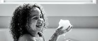 Young girl with curly hair laughing joyfully while playing with soap bubbles during bath time in bathroom. Child enjoying water play and hygiene routine.