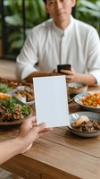 Asian man in white shirt at restaurant table with various dishes while someone holds blank white card mockup for menu or promotion display.