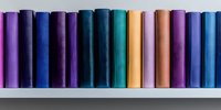 Row of colorful hardcover books standing on white shelf against neutral background, creating minimalist rainbow pattern with spines in purple, blue, teal and orange shades.