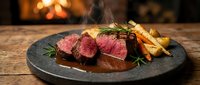 Sliced medium-rare beef steak with roasted vegetables and herbs on dark plate by cozy fireplace. Gourmet dinner presentation for restaurant menus.