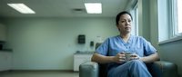 Tired Hispanic female nurse in scrubs sitting in break room holding coffee cup, looking thoughtful after long hospital shift.