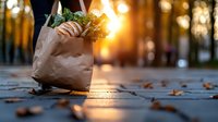Paper grocery bag filled with fresh produce and greens on cobblestone path at sunset, golden sunlight streaming through trees, fallen autumn leaves scattered around.