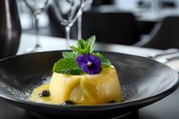 Elegant lemon dessert garnished with fresh mint leaves and purple edible flower on black ceramic plate, fine dining presentation in restaurant setting.