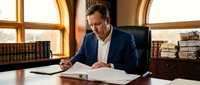 Middle-aged Caucasian businessman in navy suit reviewing documents at wooden desk in elegant office with law books and warm lighting for corporate content.