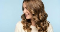 Young Caucasian woman with long wavy brown hair smiling and looking away, wearing cream sweater against light blue background for beauty and lifestyle.