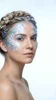 Young woman with intricate silver face paint featuring floral patterns and braided updo hairstyle against white background for fantasy makeup concepts.