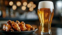 Cold glass of light beer with foam and condensation drops served with spicy buffalo chicken wings on dark plate against blurred bar background.