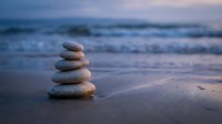 Balanced stone stack on sandy beach at sunset with gentle ocean waves creating peaceful zen meditation atmosphere for wellness and mindfulness concepts.