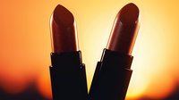 Two dark silhouettes of lipsticks against warm orange sunset background create dramatic contrast. Close up macro shot of cosmetic products with backlight effect.
