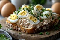 Fresh tuna salad sandwich on rustic bread topped with sliced hard-boiled eggs, chopped parsley and herbs. Close-up view on wooden cutting board with natural lighting.