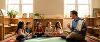Male teacher of Caucasian ethnicity reading to diverse group of young children sitting on floor in bright classroom with wooden shelves and natural light from windows, suitable for education, learning, and childcare concepts.