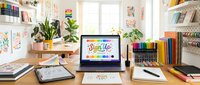 Colorful creative workspace with laptop, markers, sketchbooks, and art supplies on a bright desk surrounded by vibrant artwork prints.