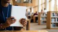 Young adult male of African descent holding blank spiral notebook in library setting, wearing blue lanyard and dark shirt, suitable for education, learning, and student concepts.