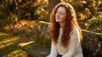 Young redhead woman with curly hair sitting peacefully on stone bench in autumn park during golden hour sunset light enjoying moment of tranquility and relaxation.
