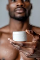 Close-up of masculine hands holding white cosmetic cream jar against blurred torso background. Soft focus portrait highlighting skincare and male grooming concept.