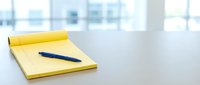 Yellow notepad with blue pen on white desk, office workspace with bright window background, business stationery for writing and planning.