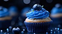 Fresh blueberry cupcake with swirled blue buttercream frosting and fresh berry on top against dark background with bokeh lights and water droplets.