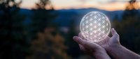 Hands holding glowing flower of life sacred geometry symbol at sunset with blurred natural landscape background for spiritual wellness meditation concepts.