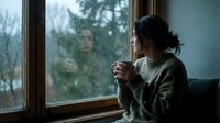 Woman with dark hair in bun, wearing cozy sweater, holding warm beverage while gazing out rainy window. Moody, contemplative atmosphere for lifestyle and wellness content.