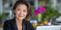 Confident Asian businesswoman with warm smile in home office environment with laptop and colorful flowers in background.