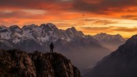 Silhouette of hiker standing on rocky mountain peak at dramatic sunset with golden sky, snow-capped peaks and flying birds in misty valley below.