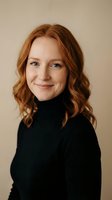 Professional portrait of confident redhead woman in black turtleneck sweater smiling warmly at camera against neutral beige background for business headshots.