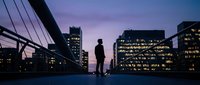 Silhouette of businessman standing on modern bridge at twilight with illuminated office buildings and purple sky creating urban evening atmosphere.