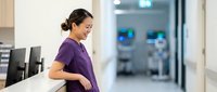 Pregnant woman with dark hair, Southeast Asian ethnicity, wearing purple shirt, standing in modern office or medical clinic environment, holding belly, expecting motherhood concept.