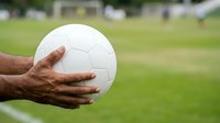 Male athlete with dark skin tone holding white soccer ball on grass field during daytime outdoor sports training or match preparation session.