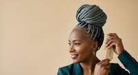Smiling African woman with gray braided hair styled in updo bun, wearing dark blazer and hoop earrings against neutral beige background. Professional portrait for business, beauty, and lifestyle content.