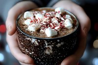 Cozy hands holding mug of hot chocolate topped with mini marshmallows and chocolate shavings, warm winter drink against blurred festive lights background.