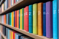 Colorful hardcover books arranged in rainbow order on wooden library shelves, creating vibrant spectrum pattern. Academic and educational background concept.