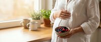 Pregnant woman in white robe holding bowl of fresh berries by sunny kitchen window. Healthy pregnancy nutrition and maternal wellness concept.