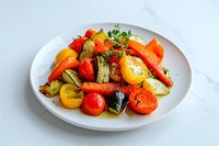 Colorful roasted vegetables with cherry tomatoes, carrots, zucchini and potatoes garnished with fresh herbs served on white ceramic plate against marble background.