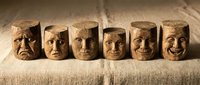 Six wooden carved face sculptures displaying a range of human emotions from sad to happy, arranged in a row on rustic linen fabric.