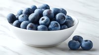 Fresh organic blueberries in white ceramic bowl on marble surface with scattered berries, healthy antioxidant superfood for breakfast and cooking.