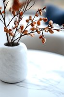 Dried brown leaves on delicate branches in white marble vase against soft blurred background, creating minimalist autumn home decor composition with natural light.