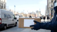 Courier in dark blue uniform and gloves holding cardboard delivery box on urban street with city buildings and vehicles in background during daytime.