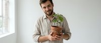 Young man with beard, Caucasian ethnicity, wearing casual beige linen shirt, holding small potted plant with green foliage indoors near window for gardening, plant care, sustainability.