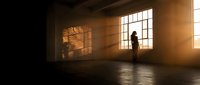 Silhouette of person standing in empty industrial warehouse with dramatic sunlight streaming through large windows creating geometric shadows on concrete floor.