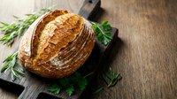 Freshly baked artisan sourdough bread loaf with golden crust on rustic wooden cutting board, garnished with fresh rosemary herbs for bakery and cooking concepts.